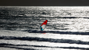 Wind Surfer contrast This landscape photograph captures a wind surfer navigating the sea as evening light reflects strongly off the water's surface. It is taken in the summer and shows the wind surfer engaged in surfing atop small waves with the sun low on the horizon, casting a silvery sheen across the ocean. The bright colors of the surf sail contrast against the darker sea, enhancing the visual impact of the wind surfer as the central subject.